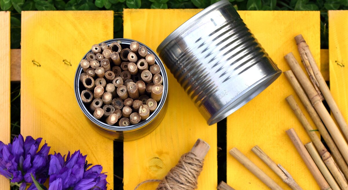 Bunte Insektenhotels in einer Blechdose, daneben eine umgestürzte Dose und Holzstäbe auf gelbem Holztisch mit lila Blumen.