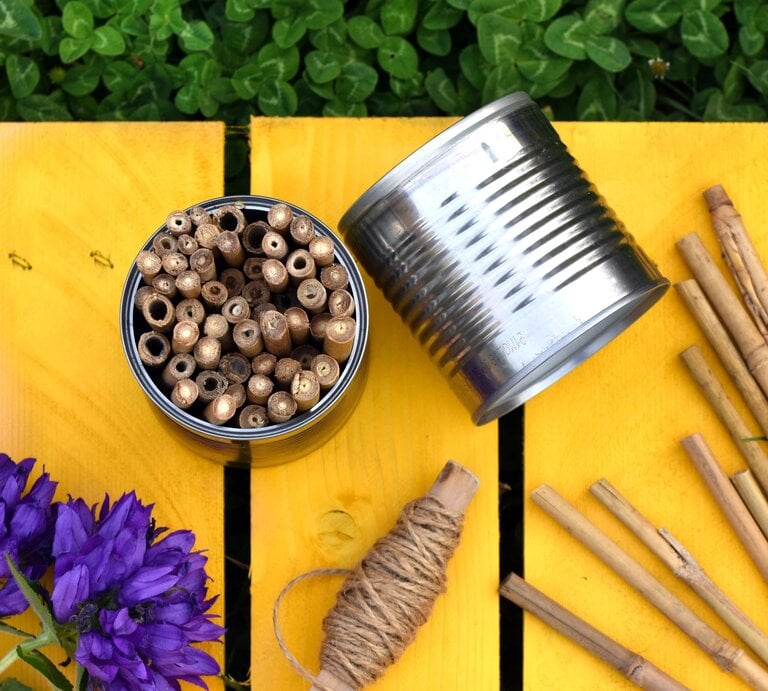 Bunte Insektenhotels in einer Blechdose, daneben eine umgestürzte Dose und Holzstäbe auf gelbem Holztisch mit lila Blumen. | © AdobeStock/Meo