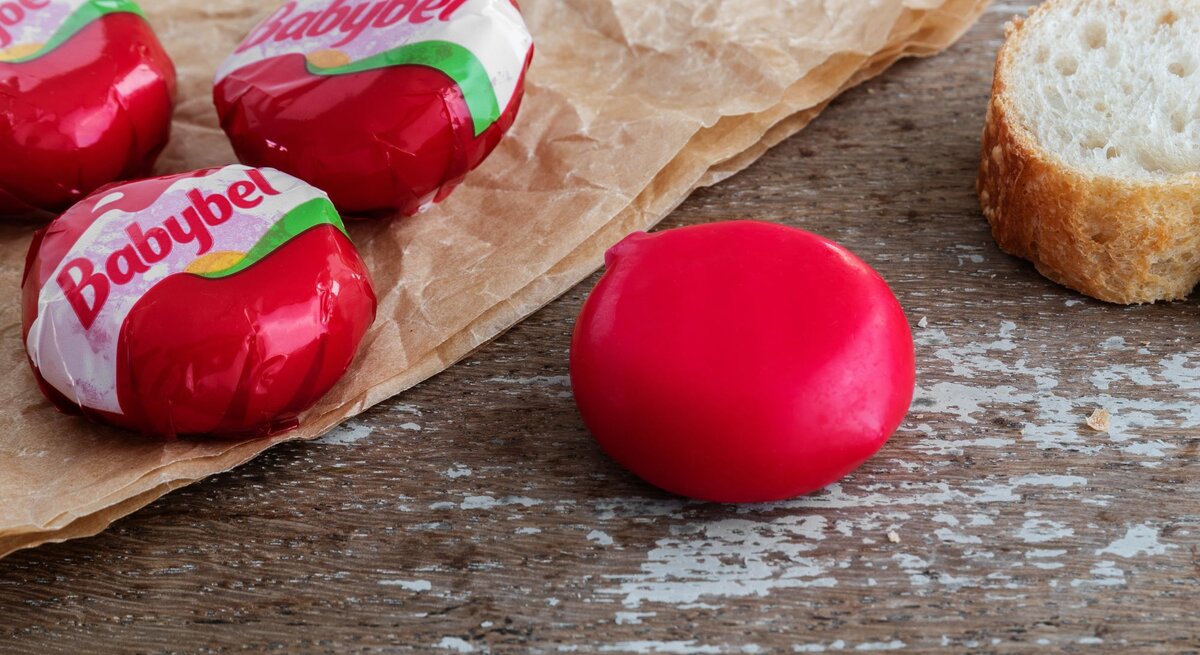 Nahaufnahme von Babybel-Käse auf rustikalem Holztisch mit Pergamentpapier und einer Baguettescheibe im Hintergrund.