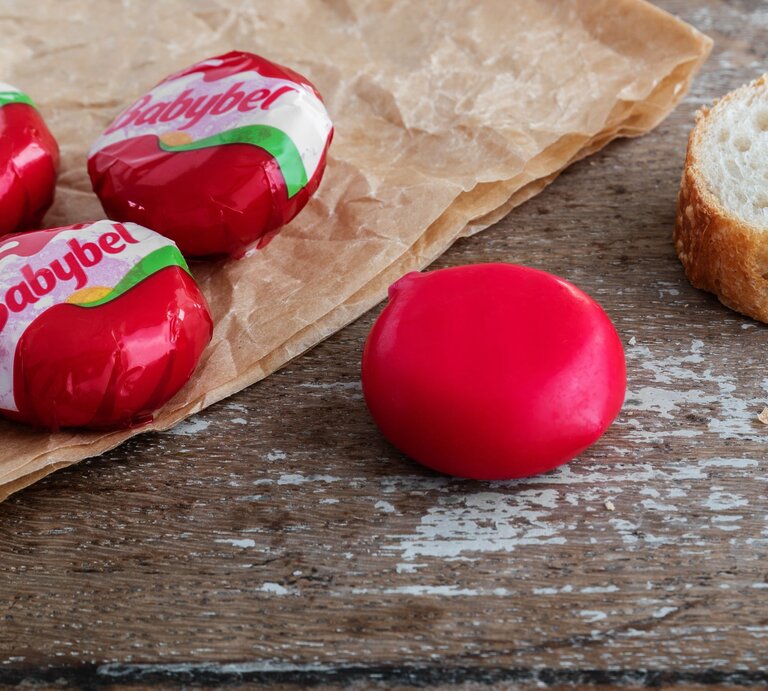 Nahaufnahme von Babybel-Käse auf rustikalem Holztisch mit Pergamentpapier und einer Baguettescheibe im Hintergrund. | © Adobe Stock/Ana Belen Garcia