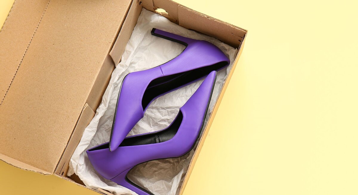 Ein Paar lila High Heels mit spitzen Absätzen liegt in einer geöffneten Pappschachtel auf gelbem Hintergrund.