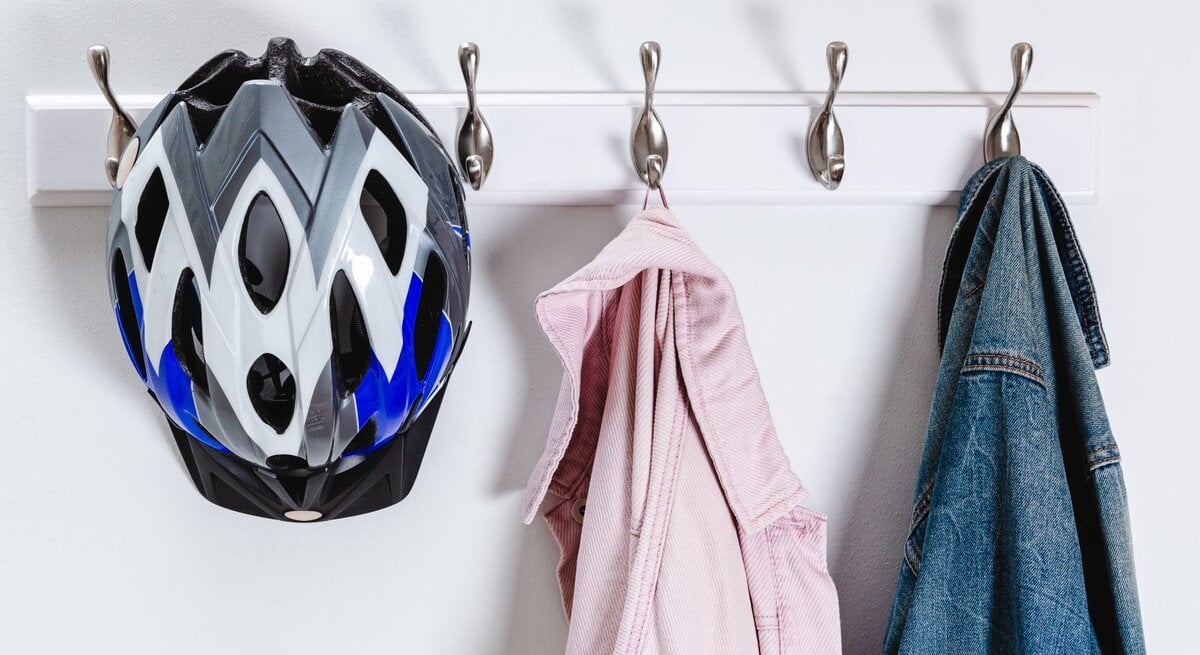 Ein Fahrradhelm mit schwarz-weiß-blauem Design hängt an einem Haken einer weißen Wandhalterung mit insgesamt fünf Haken. Neben dem Helm hängt auf der linken Seite ein rosafarbenes Hemd und auf der rechten Seite eine blaue Jeansjacke. Die Gegenstände werfen ihre Schatten an die weiße Wand, was auf eine Lichtquelle vor oder über ihnen hindeutet. Der Hintergrund ist minimalistisch und sauber, was den Fokus auf die aufgehängten Gegenstände lenkt.