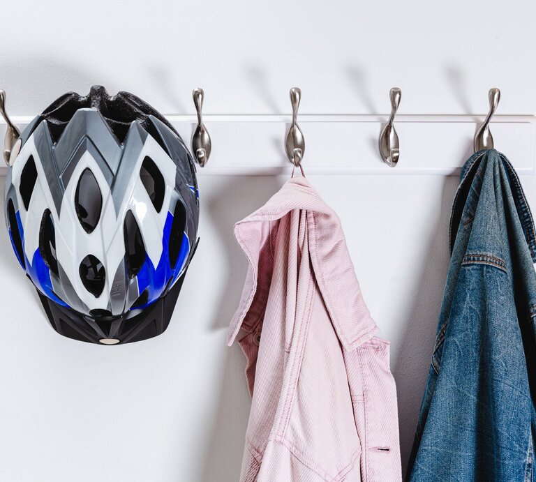 Ein Fahrradhelm mit schwarz-weiß-blauem Design hängt an einem Haken einer weißen Wandhalterung mit insgesamt fünf Haken. Neben dem Helm hängt auf der linken Seite ein rosafarbenes Hemd und auf der rechten Seite eine blaue Jeansjacke. Die Gegenstände werfen ihre Schatten an die weiße Wand, was auf eine Lichtquelle vor oder über ihnen hindeutet. Der Hintergrund ist minimalistisch und sauber, was den Fokus auf die aufgehängten Gegenstände lenkt. | © Getty Images/Alex Walker