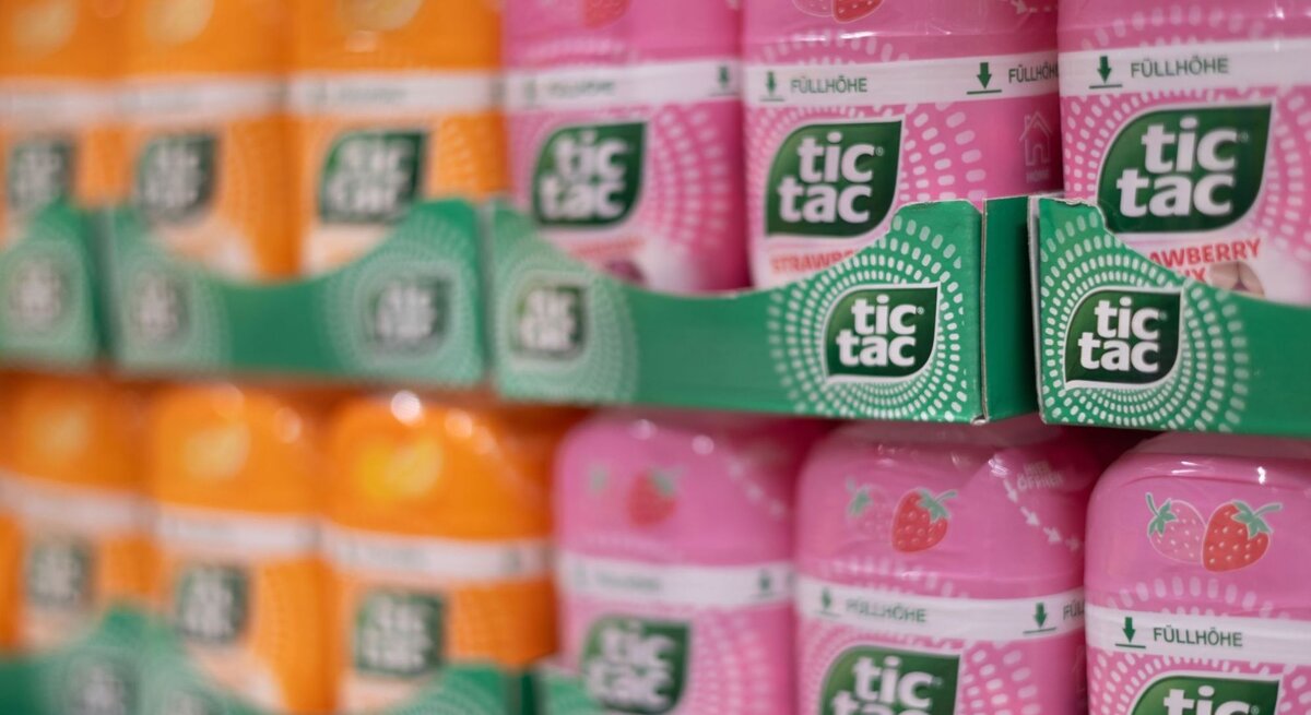 Regal mit orangefarbenen und rosa Tic Tac-Packungen in stapelbaren Pappträgern, im Vordergrund scharf fokussiert, im Hintergrund leicht verschwommen.