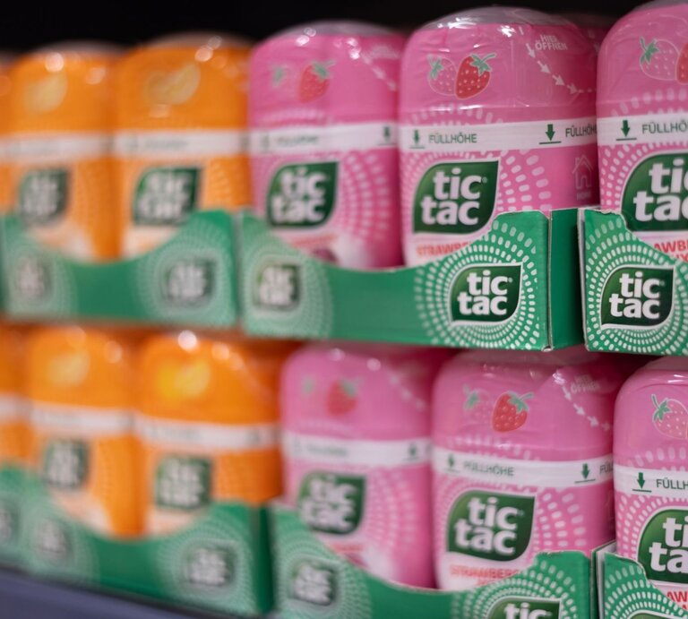 Regal mit orangefarbenen und rosa Tic Tac-Packungen in stapelbaren Pappträgern, im Vordergrund scharf fokussiert, im Hintergrund leicht verschwommen. | © Adobe Stock/ 4kclips