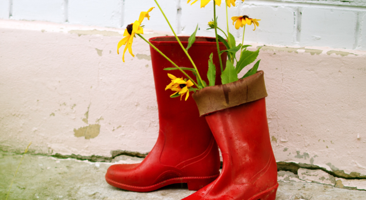 Zwei gelbe Gummistiefel mit pinken Blumen auf Pflastersteinen, daneben rote Stiefel mit Sonnenhutblüten vor heller Wand.
