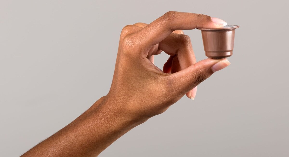  Ein Bild einer Hand, die eine gebrauchte Kaffeekapsel zwischen Daumen und Zeigefinger hält. Der Hintergrund ist einfarbig und neutral, um den Fokus auf die Kapsel zu lenken.