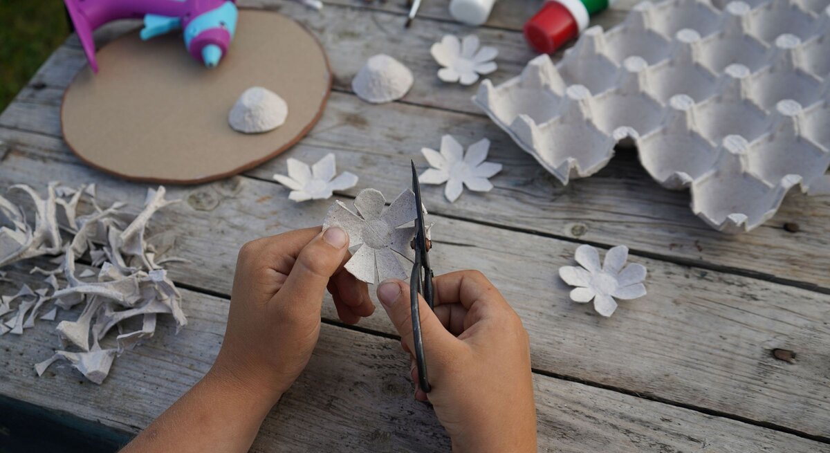 Kinderhände schneiden aus grauer Eierkartonpappe eine Blume, umgeben von Bastelmaterialien auf einem Holztisch.