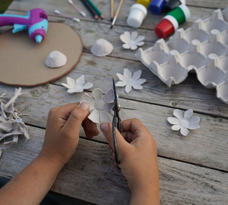 Kinderhände schneiden aus grauer Eierkartonpappe eine Blume, umgeben von Bastelmaterialien auf einem Holztisch. | © Shutterstock/ Irina Starikova1811