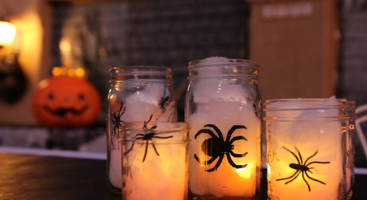 Vier Einmachgläser mit leuchtenden Kerzen und Spinnenmotiven stehen auf einem Tisch vor verschwommenem Halloween-Dekor.
