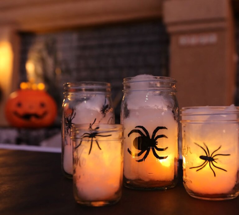Vier Einmachgläser mit leuchtenden Kerzen und Spinnenmotiven stehen auf einem Tisch vor verschwommenem Halloween-Dekor. | © Shutterstock/ Mark Breck