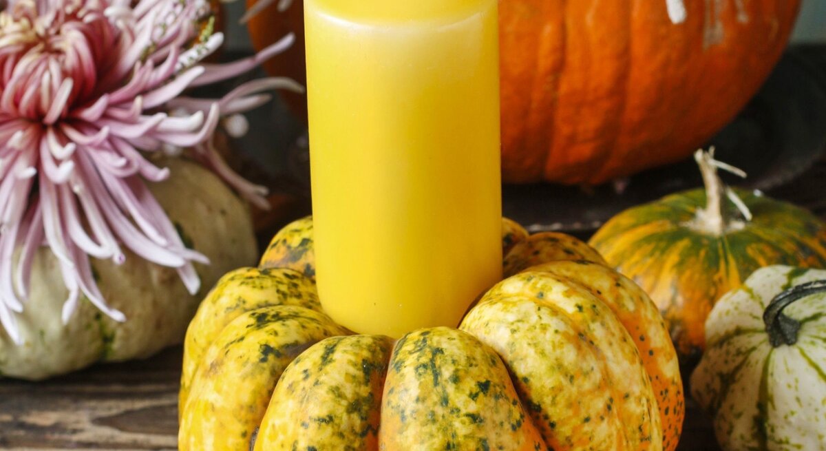 Gelbe Stumpenkerze brennt in einem dekorativen Kürbis auf rustikalem Holztisch, umgeben von Chrysanthemen und Zierkürbissen.