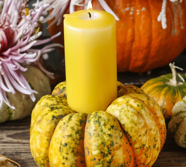 Gelbe Stumpenkerze brennt in einem dekorativen Kürbis auf rustikalem Holztisch, umgeben von Chrysanthemen und Zierkürbissen. | © Shutterstock/Agnes Kantaruk