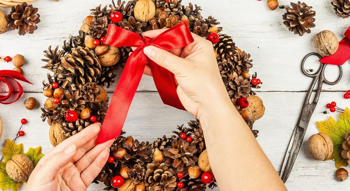 Zwei Hände binden eine rote Schleife an einen festlichen Adventskranz aus Tannenzapfen, Nüssen und roten Beeren auf hellem Holztisch.