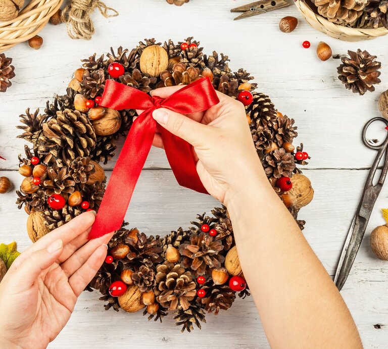 Zwei Hände binden eine rote Schleife an einen festlichen Adventskranz aus Tannenzapfen, Nüssen und roten Beeren auf hellem Holztisch. | © GettyImages/Fuzullhanum