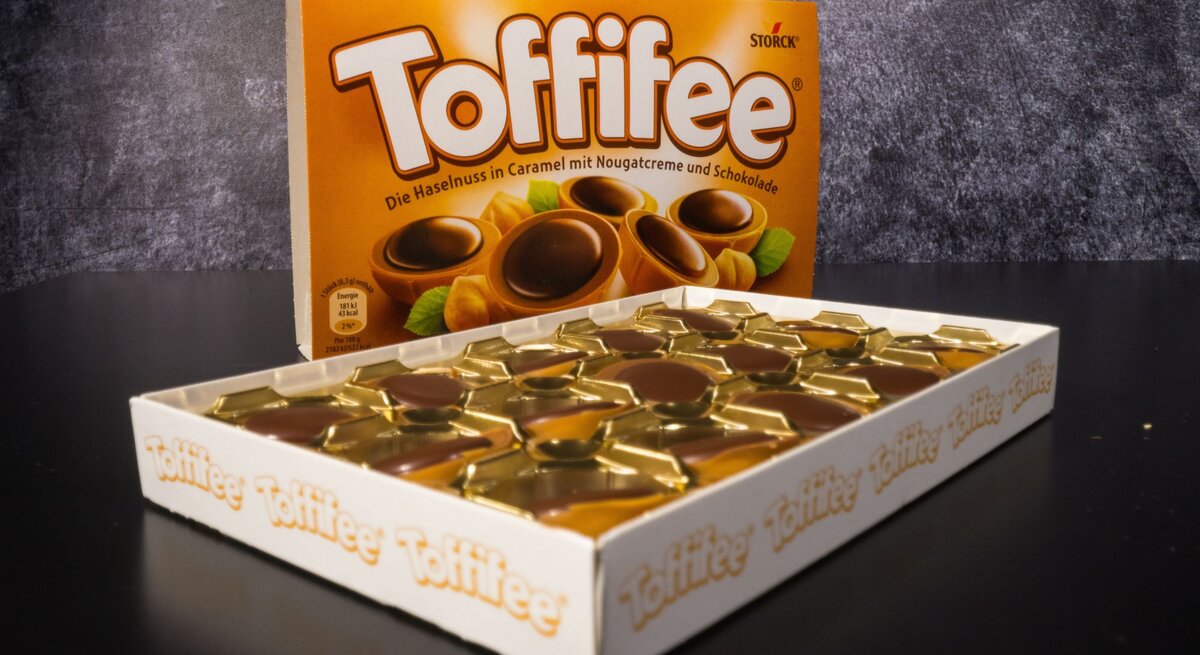Geöffnete Packung Toffifee mit goldenen Förmchen und Schokoladenpralinen, dahinter die braune Verpackung auf schwarzem Untergrund.