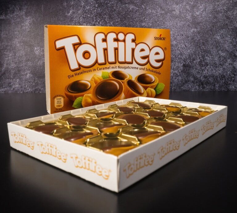 Geöffnete Packung Toffifee mit goldenen Förmchen und Schokoladenpralinen, dahinter die braune Verpackung auf schwarzem Untergrund. | © Adobe Stock/4kclips