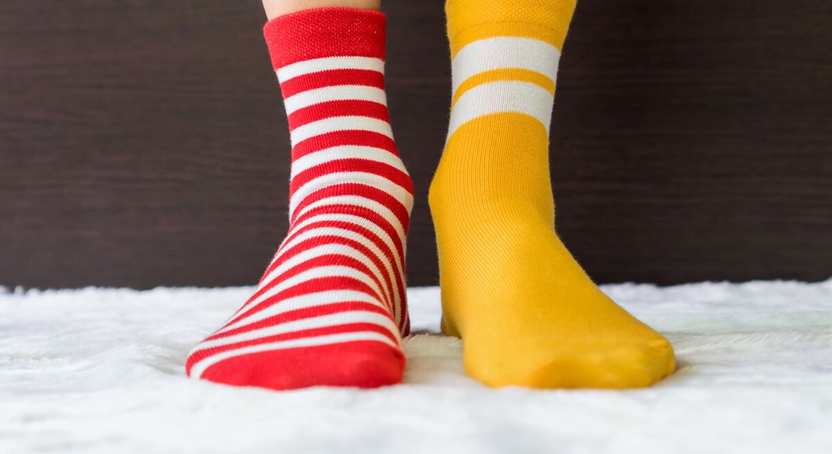 Zwei Füße in ungleichen Socken, eine rot-weiß gestreift, die andere gelb mit weißen Streifen, auf flauschigem Teppich vor dunklem Hintergrund.
