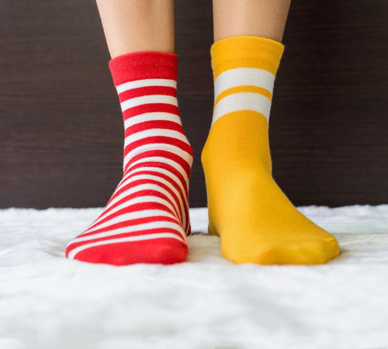 Zwei Füße in ungleichen Socken, eine rot-weiß gestreift, die andere gelb mit weißen Streifen, auf flauschigem Teppich vor dunklem Hintergrund. | © Adobe Stock/sitthiphong