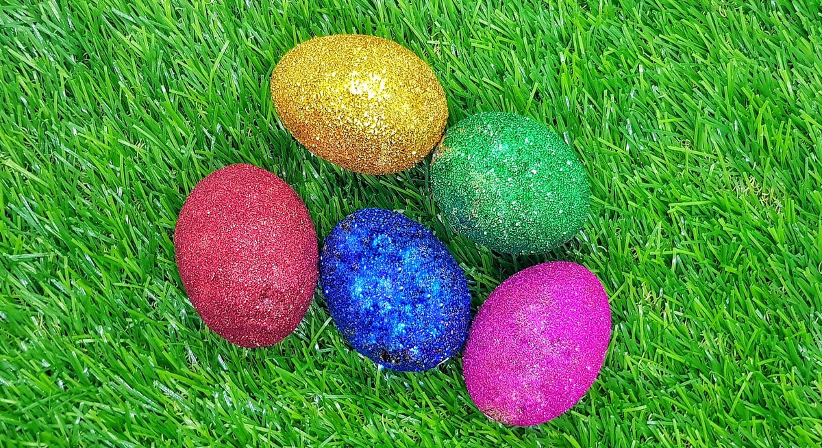 Fünf glitzernde Ostereier in Rot, Blau, Grün, Gold und Pink liegen dicht beieinander auf frischem, leuchtend grünem Gras.