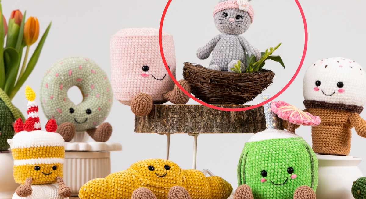Ein gehäkelter grauer Hase mit rosa Stirnband und Blüte sitzt in einem kleinen Vogelnest, umgeben von grünen Pflanzen auf einem Holzpodest.