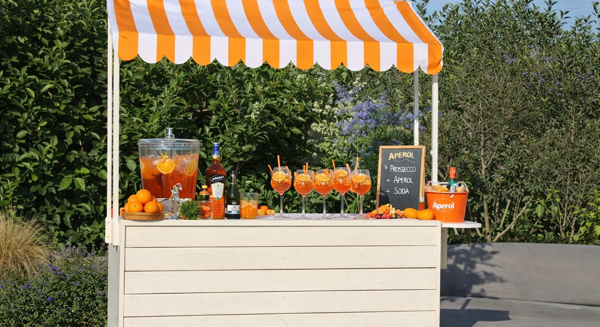 Ein mobiler Cocktailstand unter orangeweißer Markise zeigt gefüllte Aperolgläser, Spender mit Zitrusfrüchten und Deko Garten.