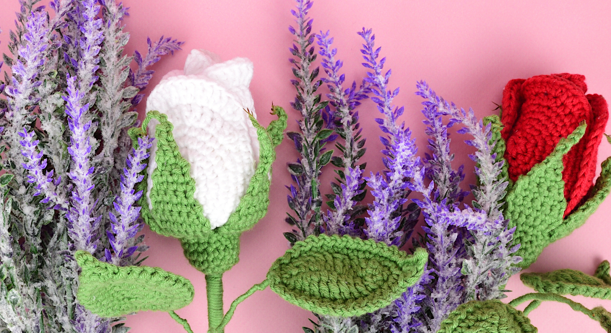 Handgehäkelte Blume mit grünen Blättern, weißen und roten Blüten vor einem Arrangement aus lilafarbenem Lavendel auf rosa Hintergrund.