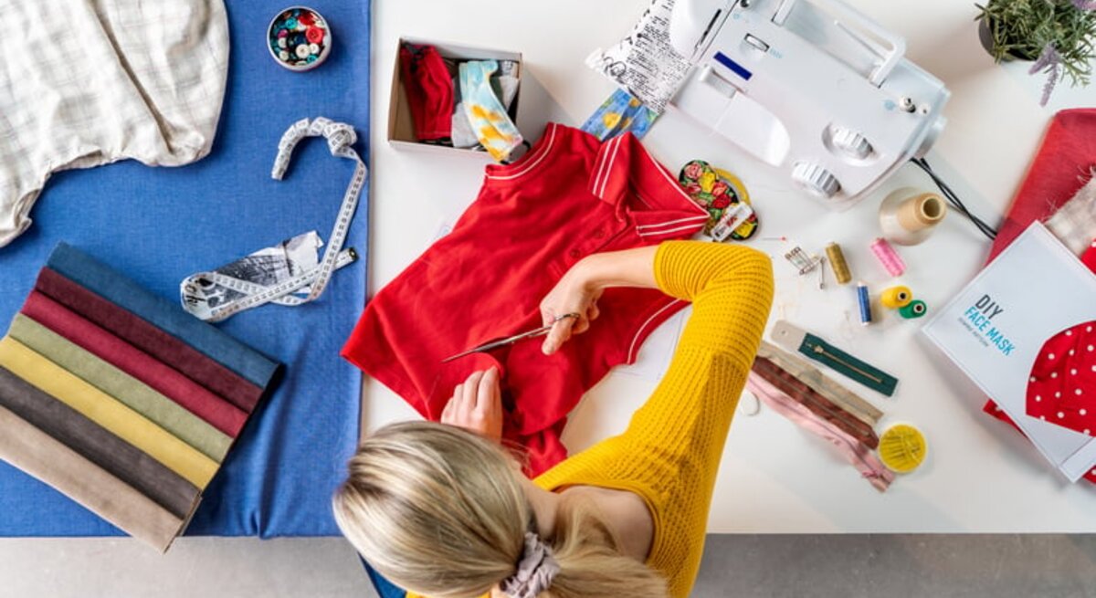 Frau schneidet altes rotes T-Shirt mit Schere um. Auf dem Tisch liegen bunte Stoffproben, Nähgarn, Knöpfe und eine Nähmaschine.