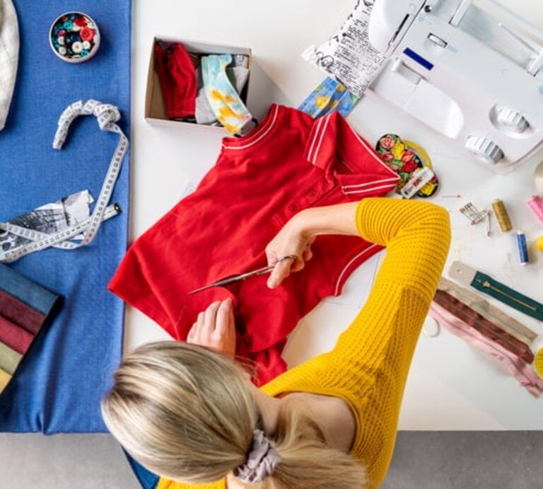 Frau schneidet altes rotes T-Shirt mit Schere um. Auf dem Tisch liegen bunte Stoffproben, Nähgarn, Knöpfe und eine Nähmaschine. | © Getty Images/ Halfpoint Images