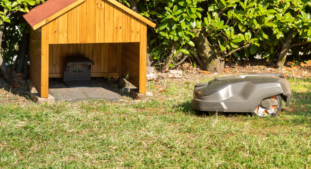 Ein Rasenmähroboter fährt bei Sonnenschein über eine Wiese in Richtung einer kleinen DIY-Garage mit Ladestation.