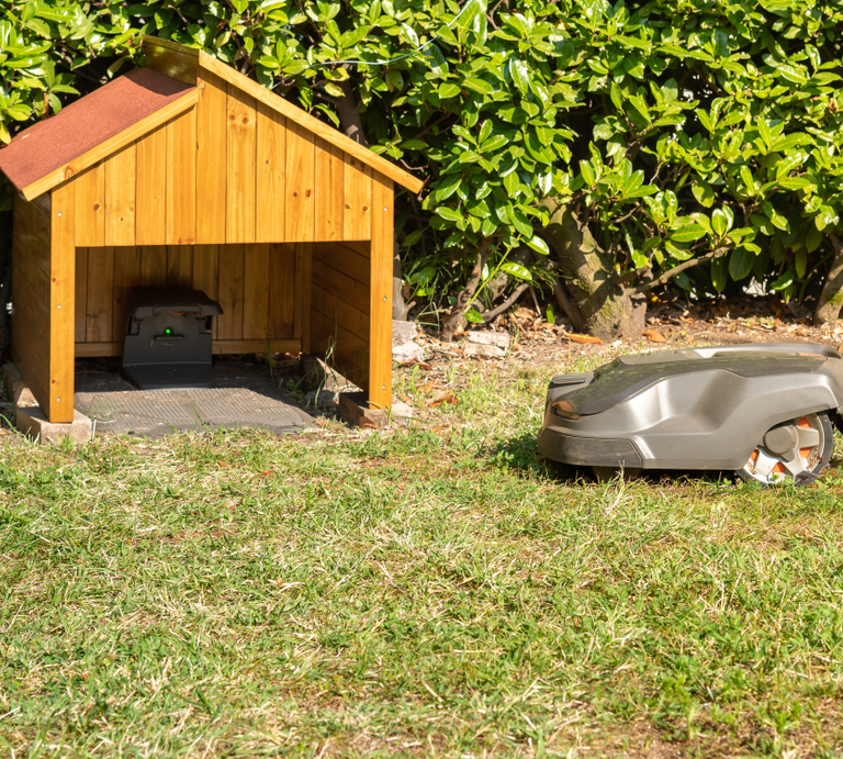 Ein Rasenmähroboter fährt bei Sonnenschein über eine Wiese in Richtung einer kleinen DIY-Garage mit Ladestation. | © AdobeStock/EleSi