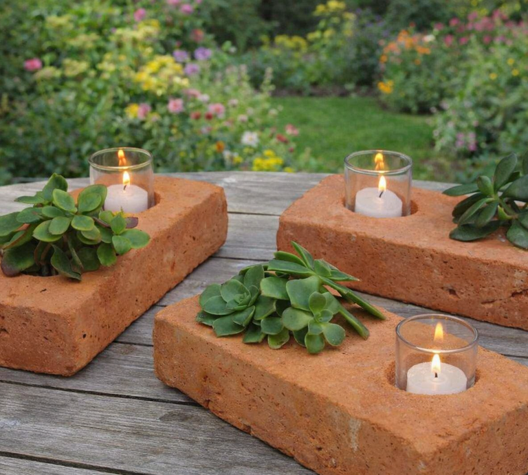 Ziegelsteine als Kerzenhalter mit Teelichtern und Sukkulenten ruhig auf Holztisch, vor üppigem Garten mit bunten Blumen. | © Erstellt mit KI (Generiert mit ChatGPT) 