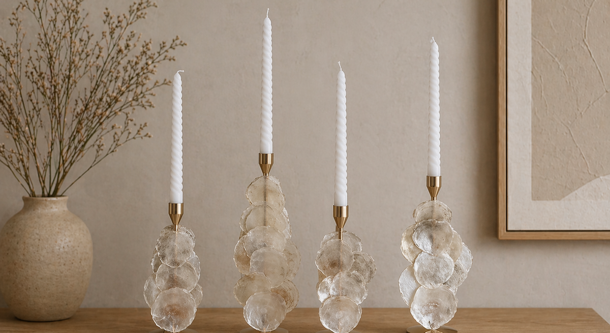 Vier elegante Kerzenständer aus Glas und Gold tragen weiße Stabkerzen auf einem Holztisch, dahinter ruhige, helle Wand und zarte Trockenpflanzen.