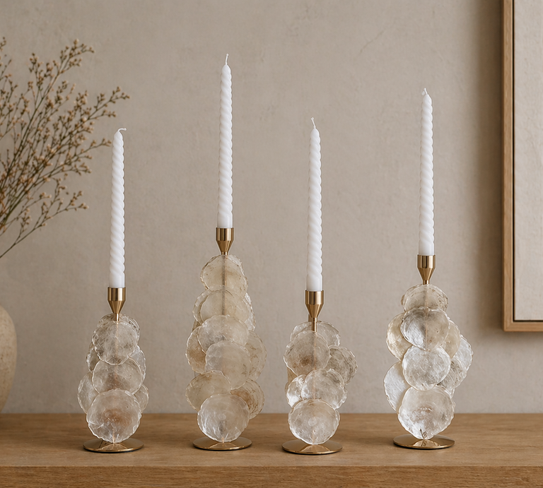 Vier elegante Kerzenständer aus Glas und Gold tragen weiße Stabkerzen auf einem Holztisch, dahinter ruhige, helle Wand und zarte Trockenpflanzen. | © Erstellt mit KI (Generiert mit ChatGPT) 
