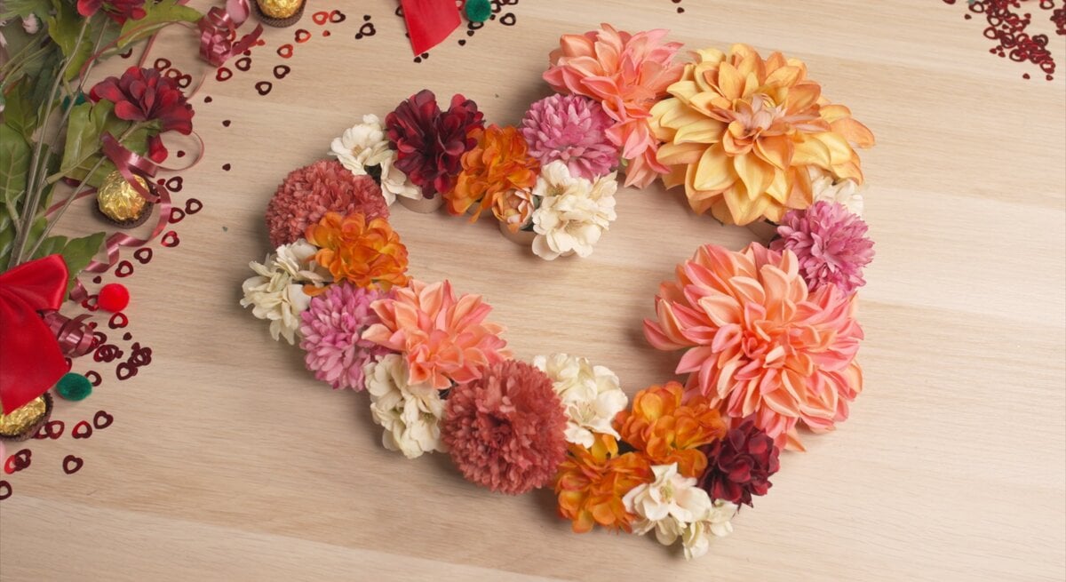 Herzförmiger Kranz aus bunten Kunstblumen in Rosa-, Orange- und Cremetönen liegt als DIY-Deko auf einem hellen Holztisch, umgeben von Streudeko und Schleifen.