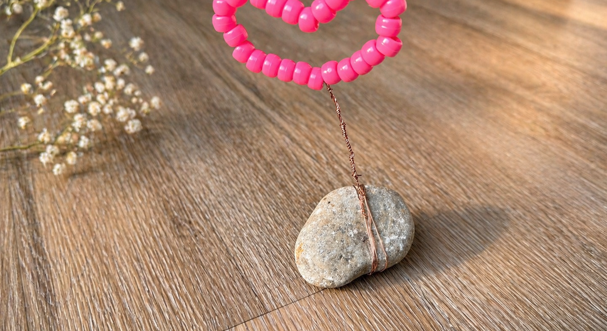 Pinkes Herz aus Perlen auf Draht ragt aus einem kleinen grauen Stein, dekorativ platziert auf warmem Holzboden mit zarten Blüten.