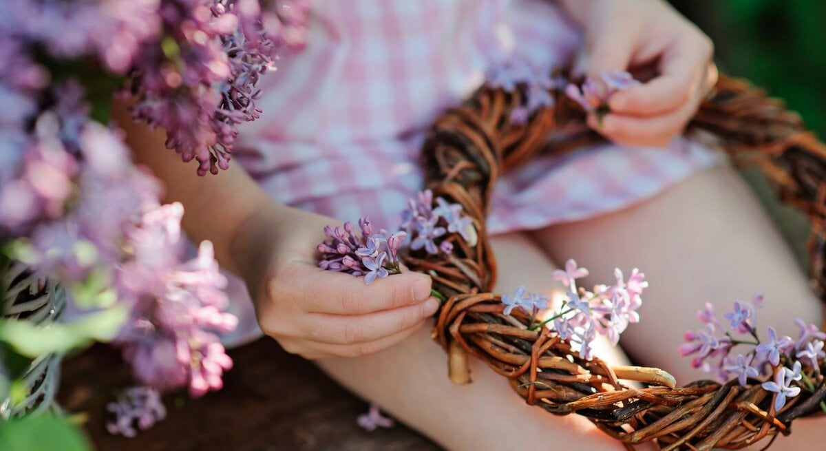 Kind bastelt einen Blumenkranz aus lila Fliederblüten im Freien. 