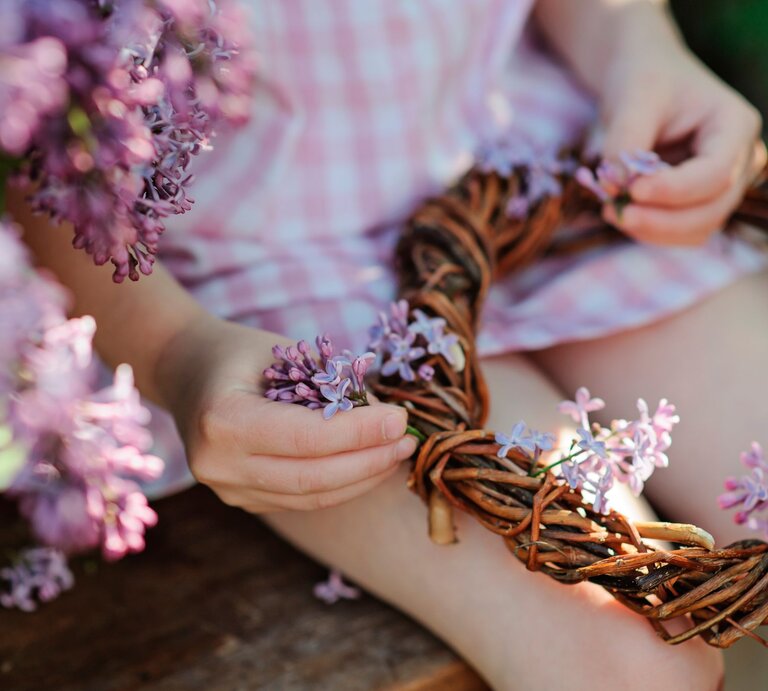 Kind bastelt einen Blumenkranz aus lila Fliederblüten im Freien.  | © Shutterstock/Maria Evseyeva
