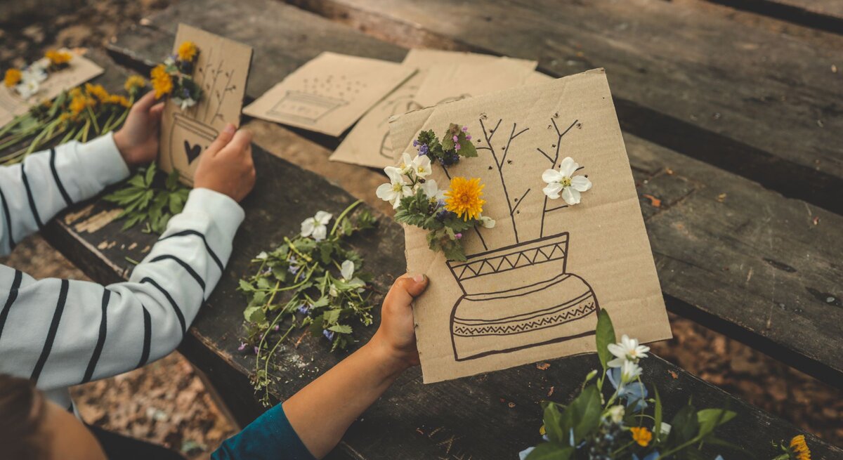 Kinderhände gestalten auf einem Holztisch Karten mit Löwenzahn, Gänseblümchen und Blumen und gezeichneten Vasen aus braunem Karton. | © GettyImages/Bohdan Bevz