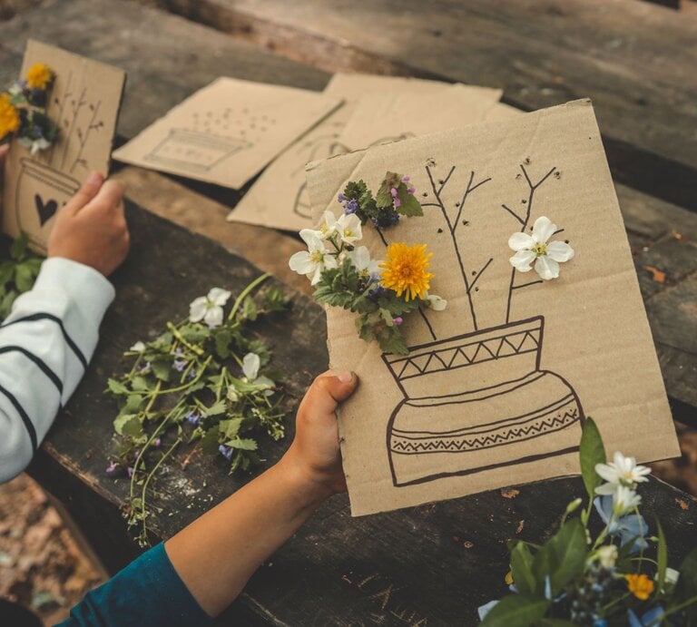Kinderhände gestalten auf einem Holztisch Karten mit Löwenzahn, Gänseblümchen und Blumen und gezeichneten Vasen aus braunem Karton. | © GettyImages/Bohdan Bevz