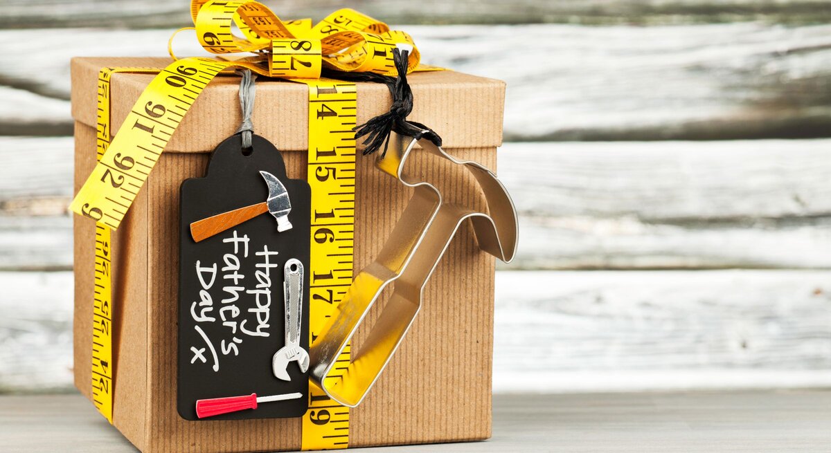 Ein braun verpacktes Geschenk mit gelbem Maßband und Werkzeug-Anhängern steht vor helzem Holzhintergrund.