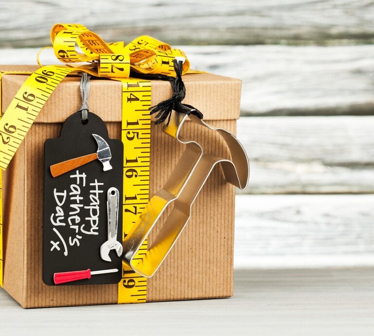 Ein braun verpacktes Geschenk mit gelbem Maßband und Werkzeug-Anhängern steht vor helzem Holzhintergrund. | © GettyImages/CatLane