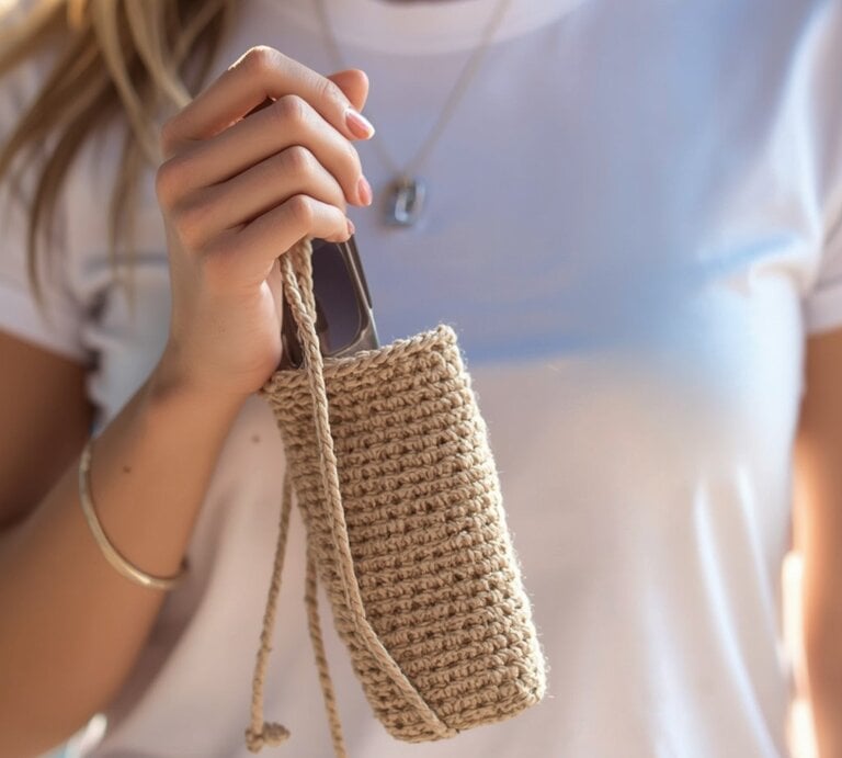 Junge Frau mit blonden Haaren hält ein handgemachtes beiges Häkel-Etui mit Sonnenbrille vor weißem T-Shirt. | © Canva / AI