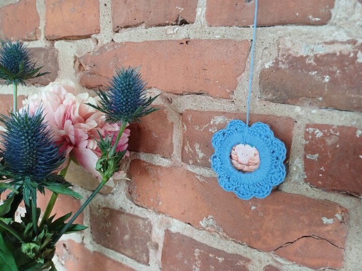 Ein Strauß rosa Pfingstrosen mit blauen Disteln steht vor einer rustikalen Ziegelwand, daneben hängt ein gehäkelter blauer Mini-Bilderrahmen. | © privat