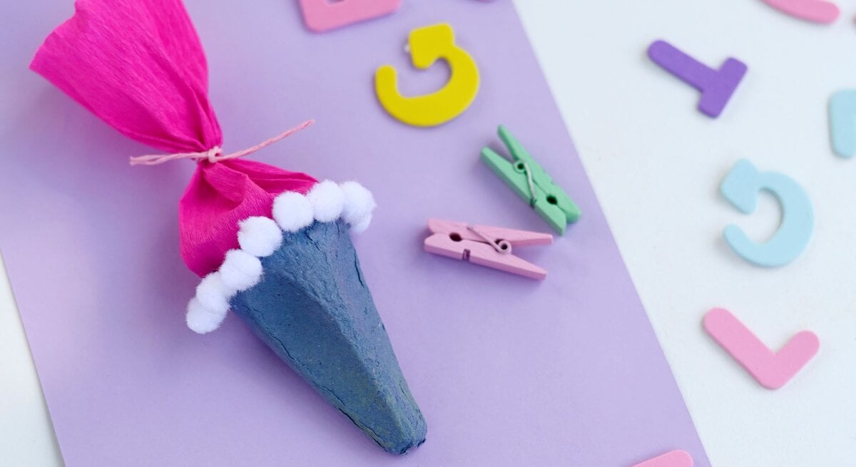 Bunte Schultüte aus Eierkarton mit pinkem Papier und weißen Pompons liegt auf lilafarbenem Bastelkarton zwischen Buchstaben. | © GettyImages/Westend61