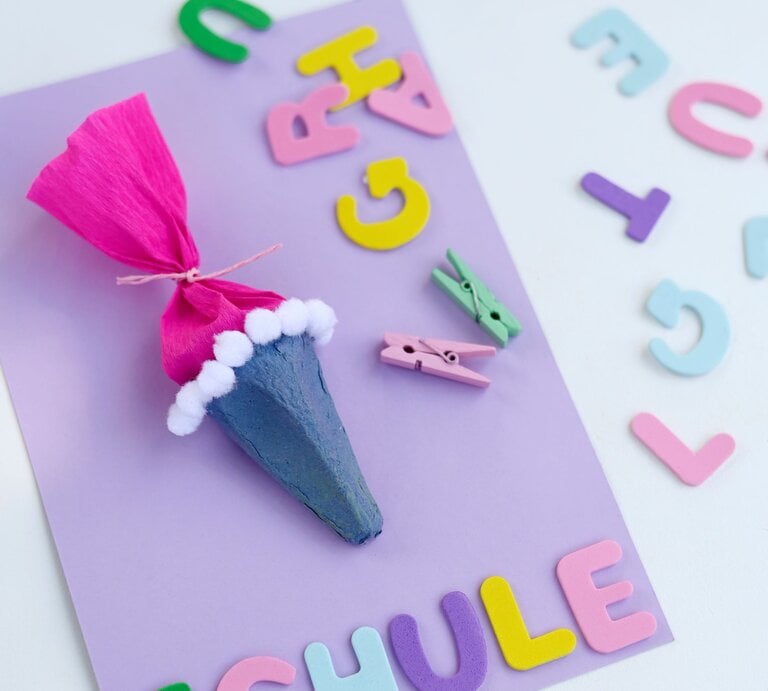 Bunte Schultüte aus Eierkarton mit pinkem Papier und weißen Pompons liegt auf lilafarbenem Bastelkarton zwischen Buchstaben. | © GettyImages/Westend61