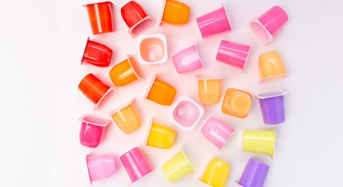 Bunte Joghurtbecher in Rosa, Gelb, Orange und Lila liegen verstreut auf weißem Hintergrund in fröhlicher Anordnung.