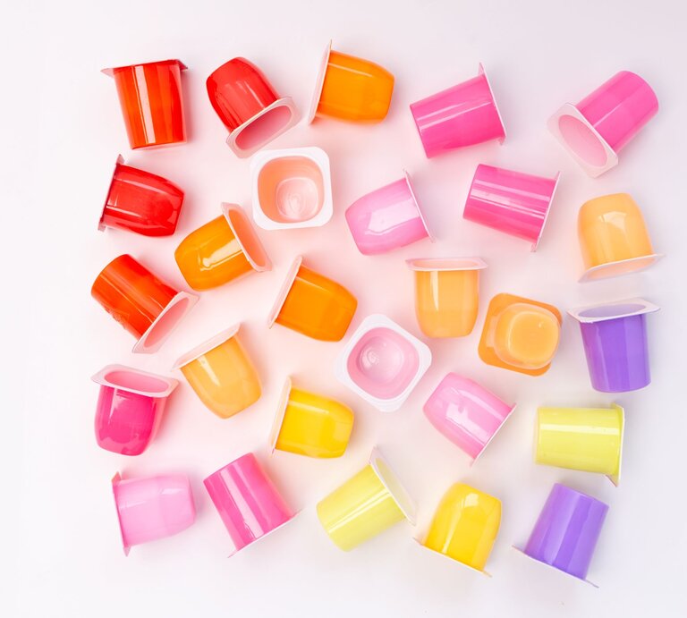 Bunte Joghurtbecher in Rosa, Gelb, Orange und Lila liegen verstreut auf weißem Hintergrund in fröhlicher Anordnung. | © Shutterstock/Elena Chevalier
