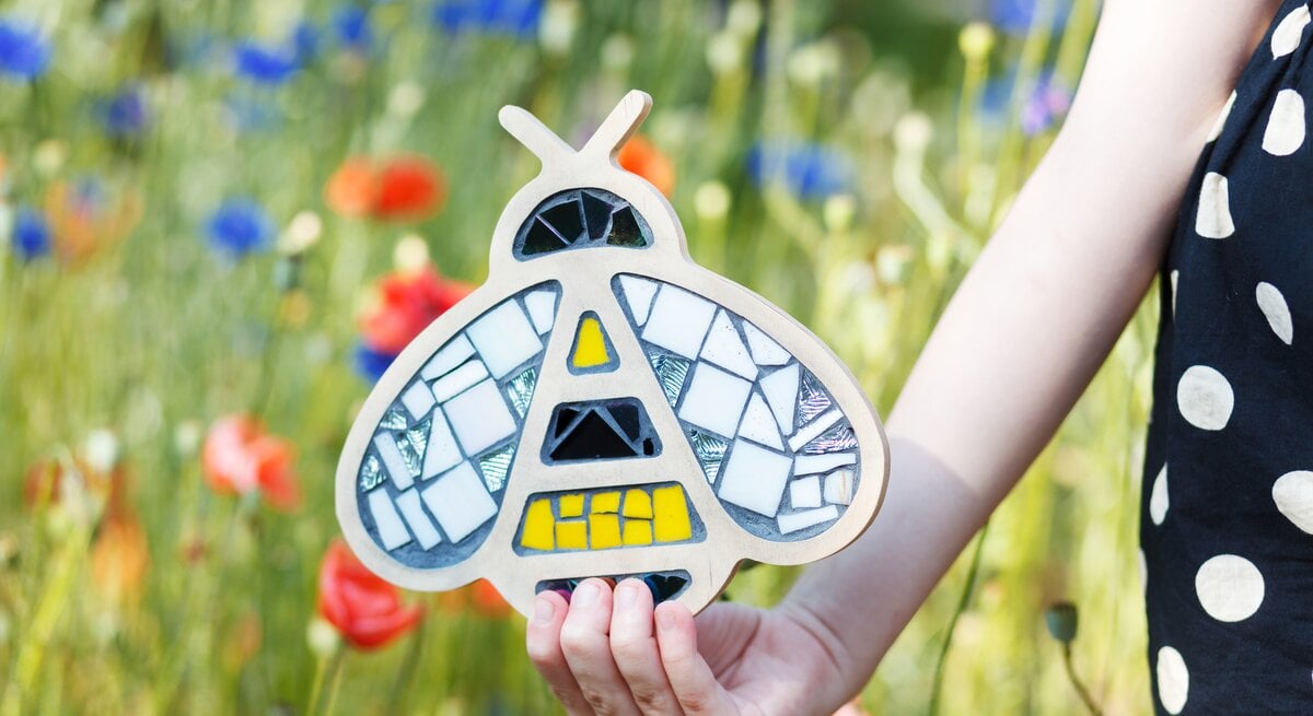 Eine Kinderhand hält eine mosaikverzierte Biene aus Holz vor einer blühenden Sommerwiese mit roten Mohnblumen und Kornblumen.