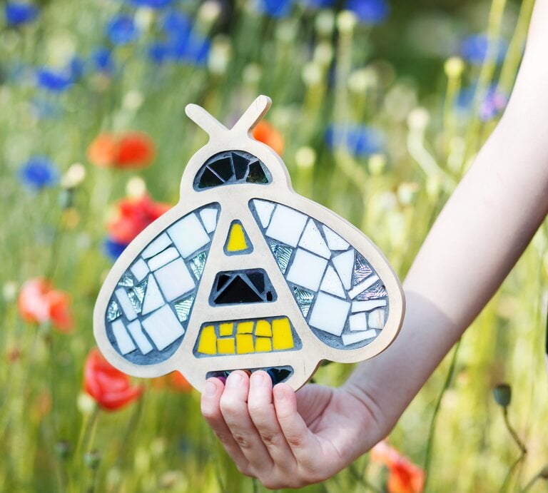Eine Kinderhand hält eine mosaikverzierte Biene aus Holz vor einer blühenden Sommerwiese mit roten Mohnblumen und Kornblumen. | © GettyImages/Natalija Grigel